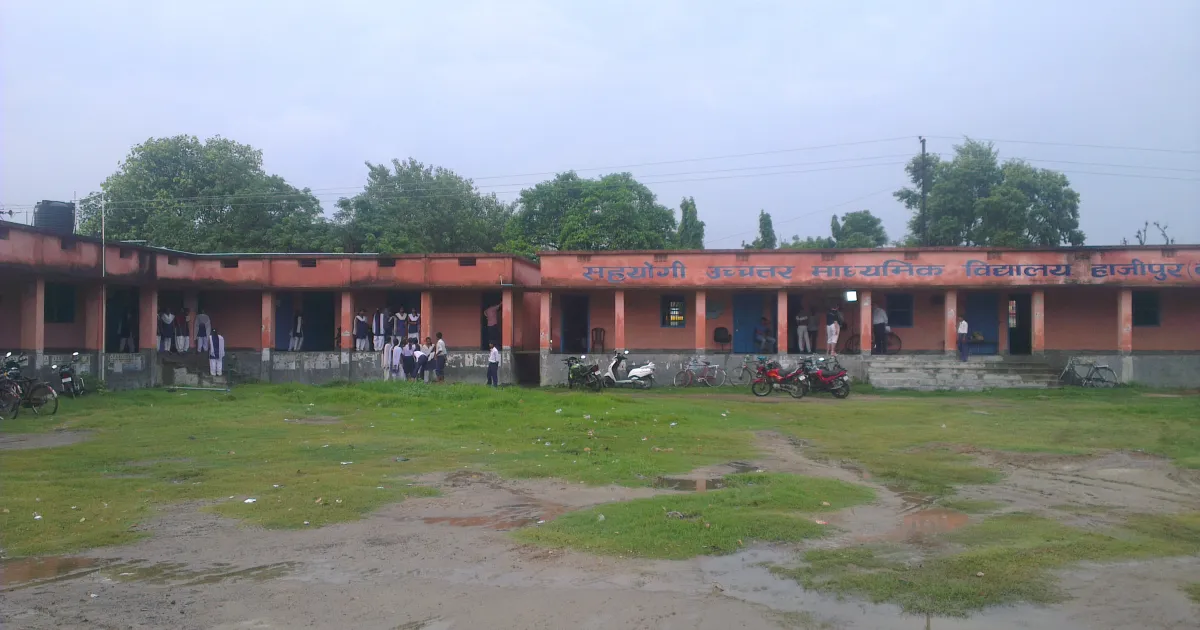 A single-storey government school building in Bihar with a red and cream wall, students on the veranda and a grassy ground in front