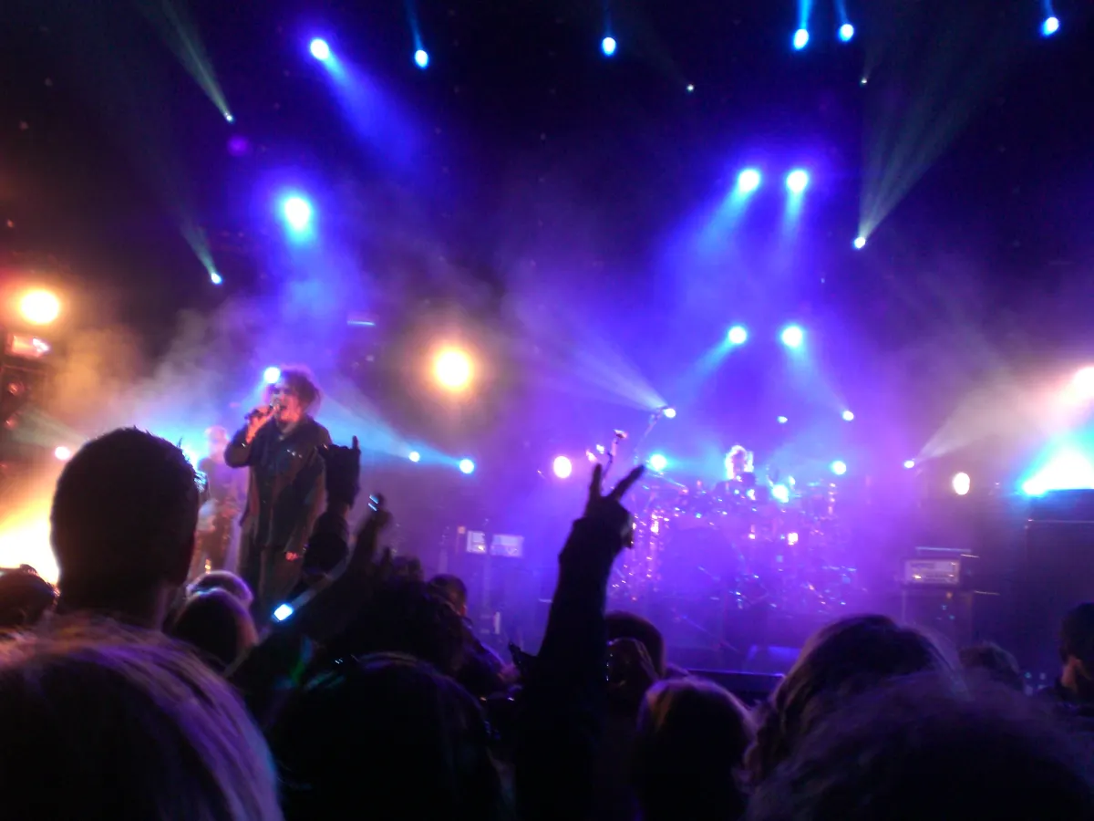 The Cure performing on a stage lit with blue and purple lights, with fans’ raised hands in the foreground