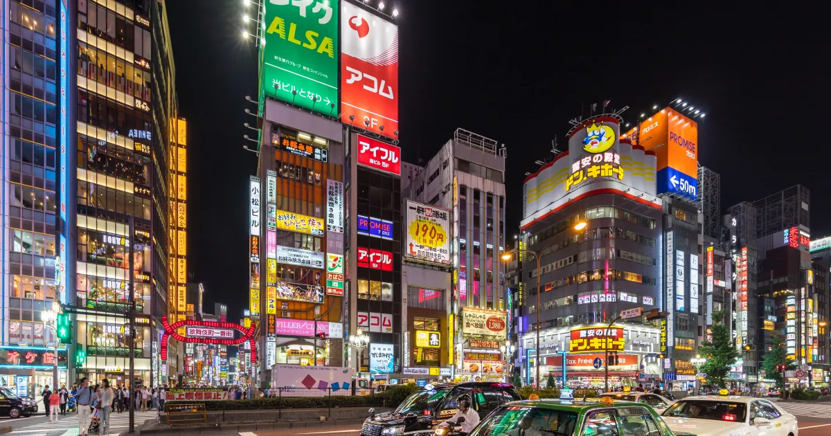 A wide avenue in Shinjuku, Tokyo at night, lined with tall buildings covered in bright neon signs and green taxis at a crossing