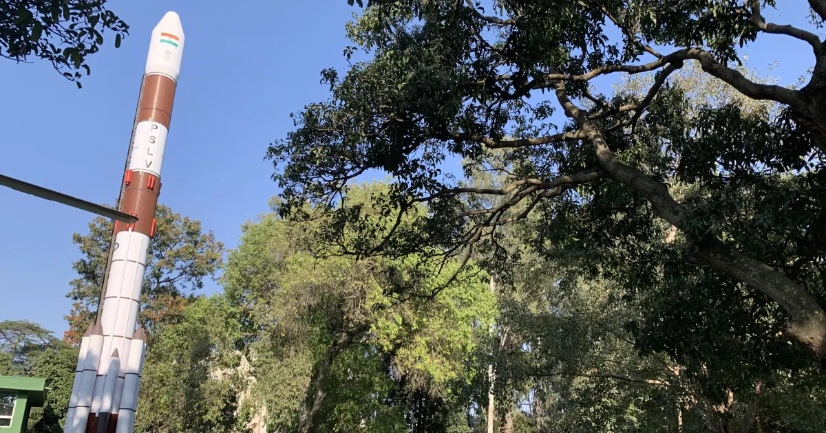 A tall white and brown PSLV rocket model standing among green trees under a clear blue sky