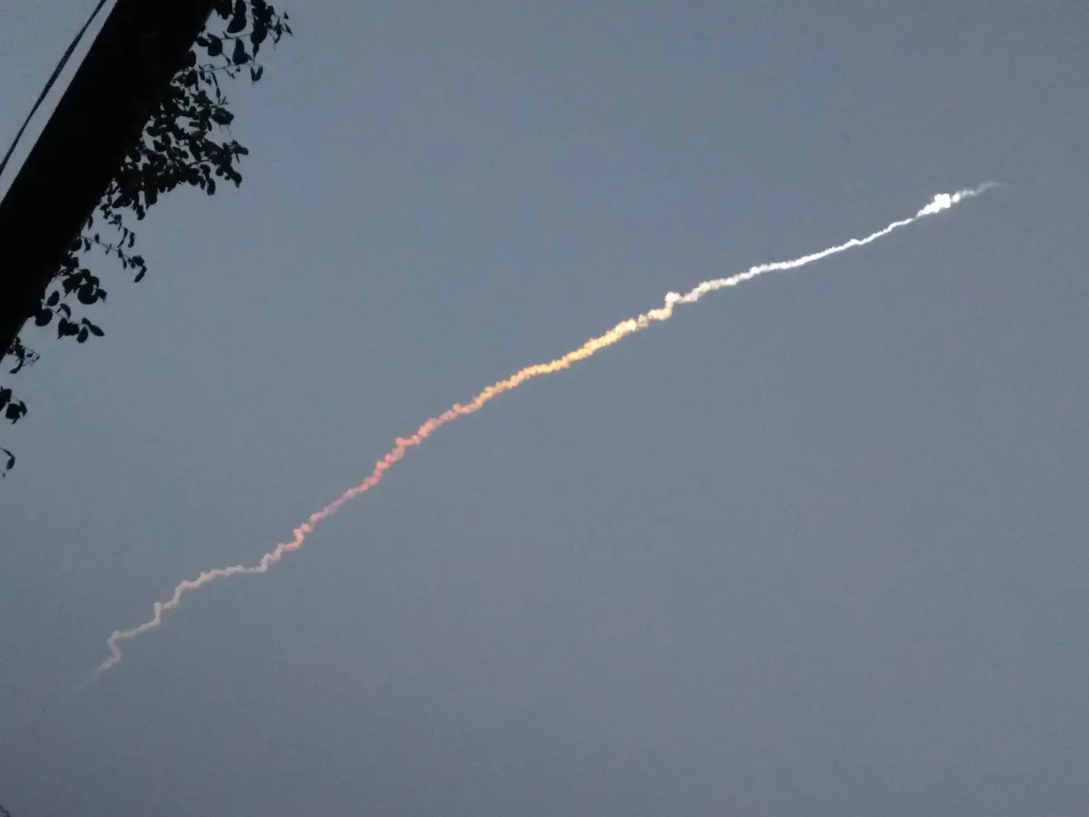 A curving white and orange smoke trail of a rocket climbing into a grey sky, seen from far away past tree branches