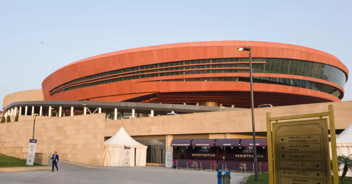Bharat Mandapam convention centre in New Delhi, a large round building with an orange-red curved roof, seen from the east entrance plaza