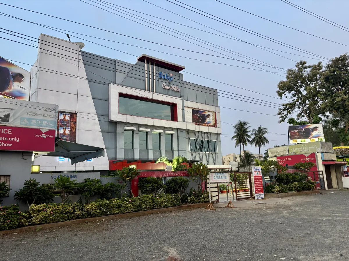A modern grey cinema building with a red-trimmed entrance, palm trees and a parking area in front