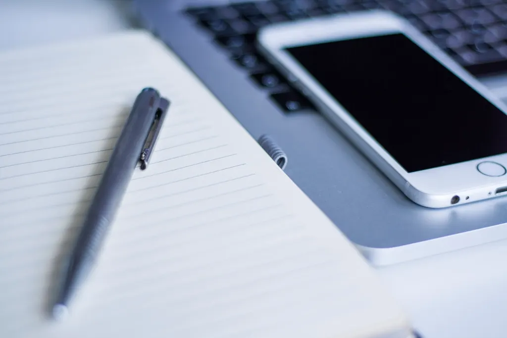 A pen on an open lined notebook next to a white smartphone and a laptop