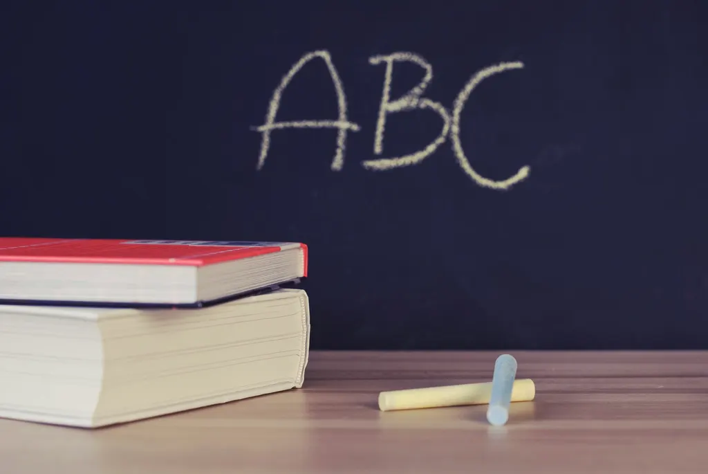 Two books and pieces of chalk on a wooden desk in front of a blackboard with ABC written in chalk