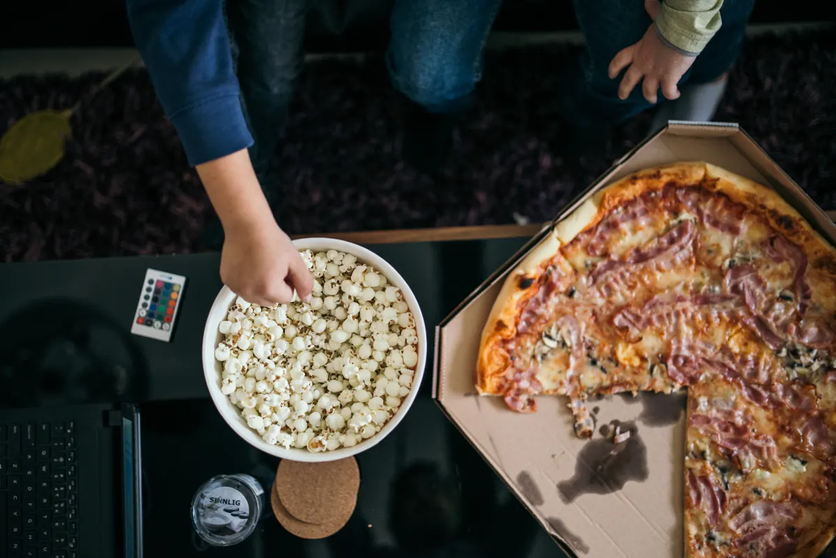 A family’s hands reaching for a bowl of popcorn and a pizza box on a table during a movie night at home