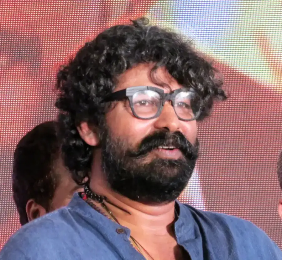 Actor Joju George with curly hair, a beard and glasses, smiling at a public event