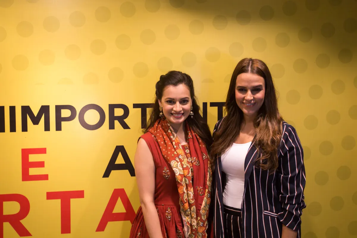 Dia Mirza and Neha Dhupia standing together in front of a yellow event backdrop