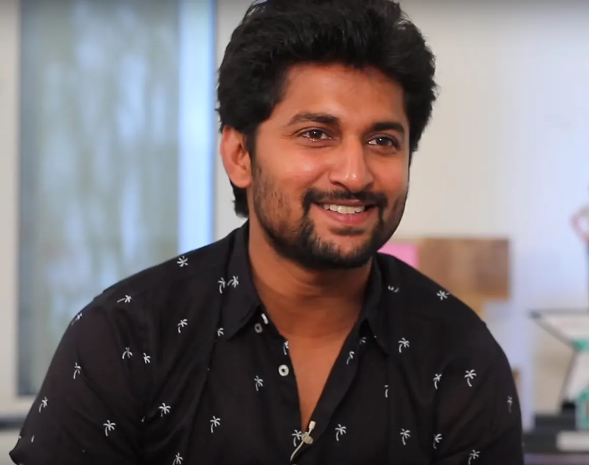 Actor Nani with a beard, smiling in a black printed shirt during an interview