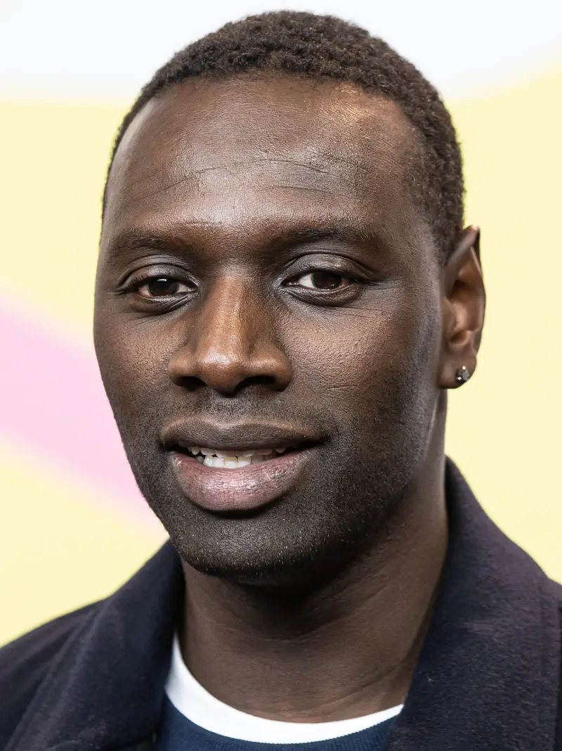 Close-up of actor Omar Sy smiling, in a dark jacket against a pale yellow background