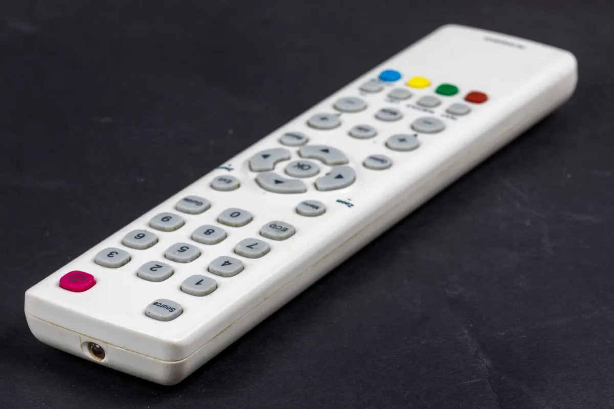 A white television remote control with coloured buttons on a dark surface