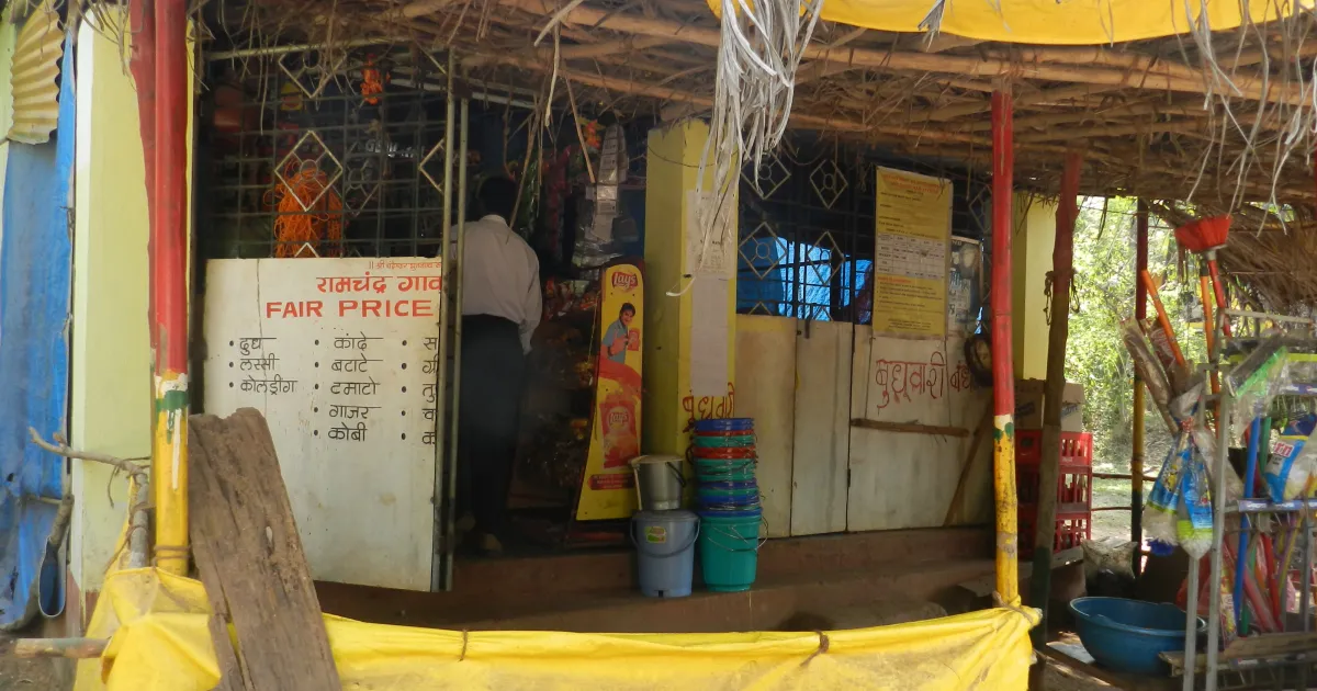 A village fair price shop in India with a hand-painted board, plastic buckets and a yellow tarpaulin in front
