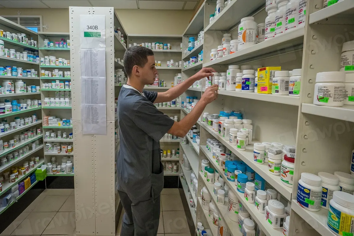 A pharmacist in a grey uniform arranging medicine bottles on tall pharmacy shelves