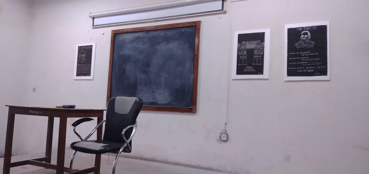 An empty classroom with a blackboard, a teacher’s table and chair, and framed Telugu posters on the wall
