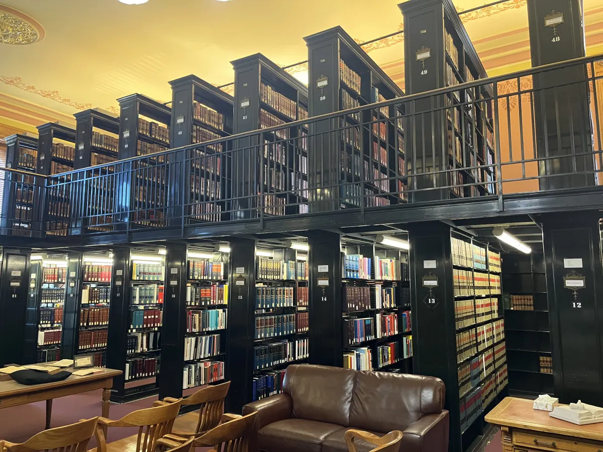 Tall dark wooden bookshelves on two levels in a library reading room, with a leather sofa and reading tables in front