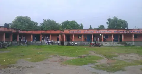 A single-storey government school building in Bihar with a red and cream wall, students on the veranda and a grassy ground in front