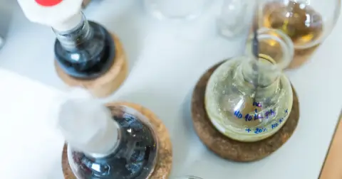 Round-bottom flasks with dark and pale yellow liquids resting on cork rings on a laboratory bench
