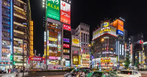 A wide avenue in Shinjuku, Tokyo at night, lined with tall buildings covered in bright neon signs and green taxis at a crossing