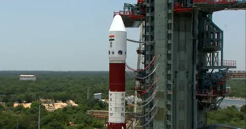 A red and white PSLV rocket standing next to its tall service tower on the launch pad at Sriharikota, with green forest behind it
