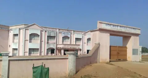 A two-storey Odisha Adarsha Vidyalaya school building behind a pink boundary wall and gate with an Odia name board