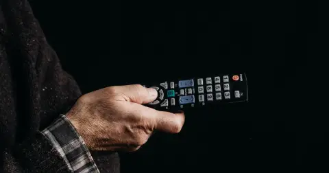 A hand in a knitted sweater holding a TV remote control against a dark background
