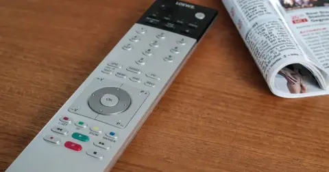 A silver television remote control lying on a wooden table next to a rolled-up magazine