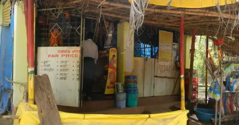 A village fair price shop in India with a hand-painted board, plastic buckets and a yellow tarpaulin in front
