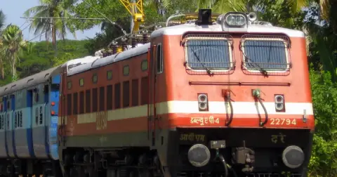 A red and white WAP-4 electric locomotive of Indian Railways pulling a passenger train past palm trees
