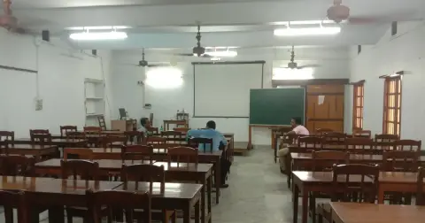 An empty college classroom with rows of wooden desks, ceiling fans and a green chalkboard at the front