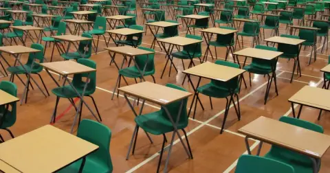 A large empty exam hall with rows of wooden-top desks and green plastic chairs on a sports floor