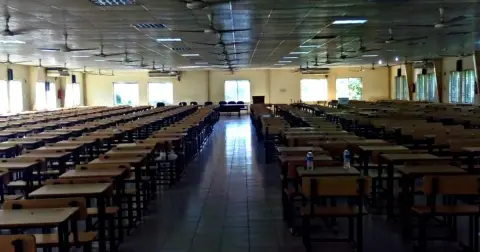 Long rows of empty wooden desks and chairs in a large examination hall with ceiling fans